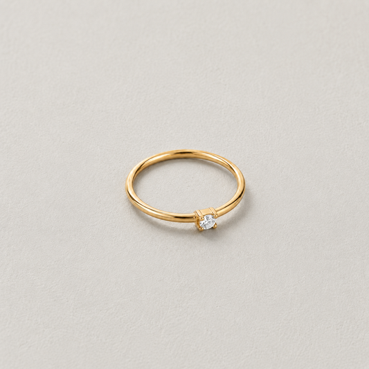 Dew Ring, smal guldfärgad ring med klar sten på ljus stenbakgrund.