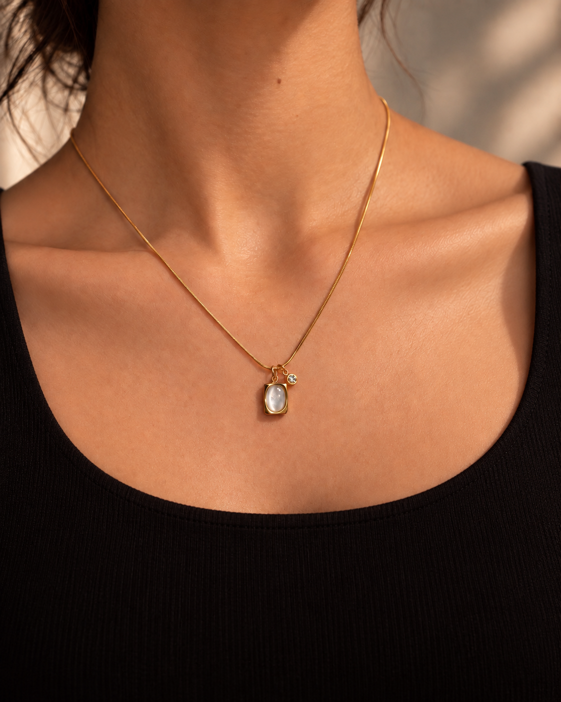 Halo Necklace i guld med oval pärldetalj och liten klar sten, buren på hals mot svart topp i varmt ljus.