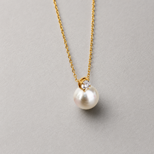 Lustre Necklace i 316 kirurgiskt stål och 18k PVD-guld, guldfärgat halsband med pärla och klar sten mot ljus neutral bakgrund.