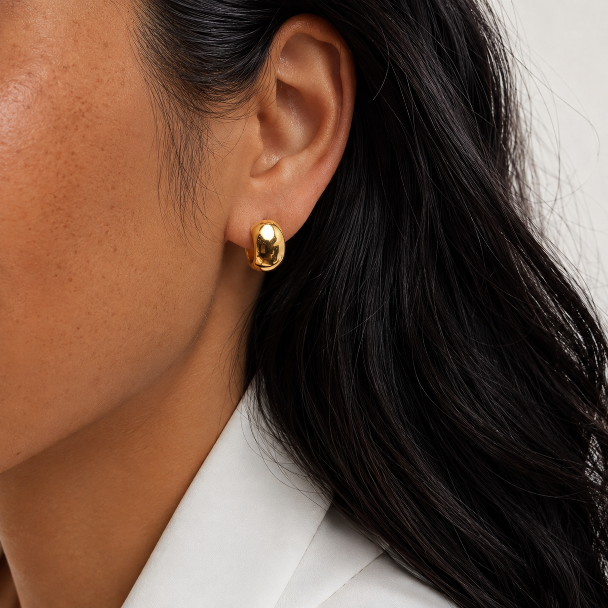 Crescent Hoop i 316 kirurgiskt stål och 18k PVD-guld, ett rundat guldfärgat hoop-örhänge buret i örat mot vit textil.