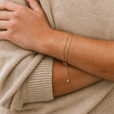 Current Bracelet i 316 kirurgiskt stål och 18k PVD-guld, ett nätt guldfärgat armband buret på handleden mot beige stickad textil.