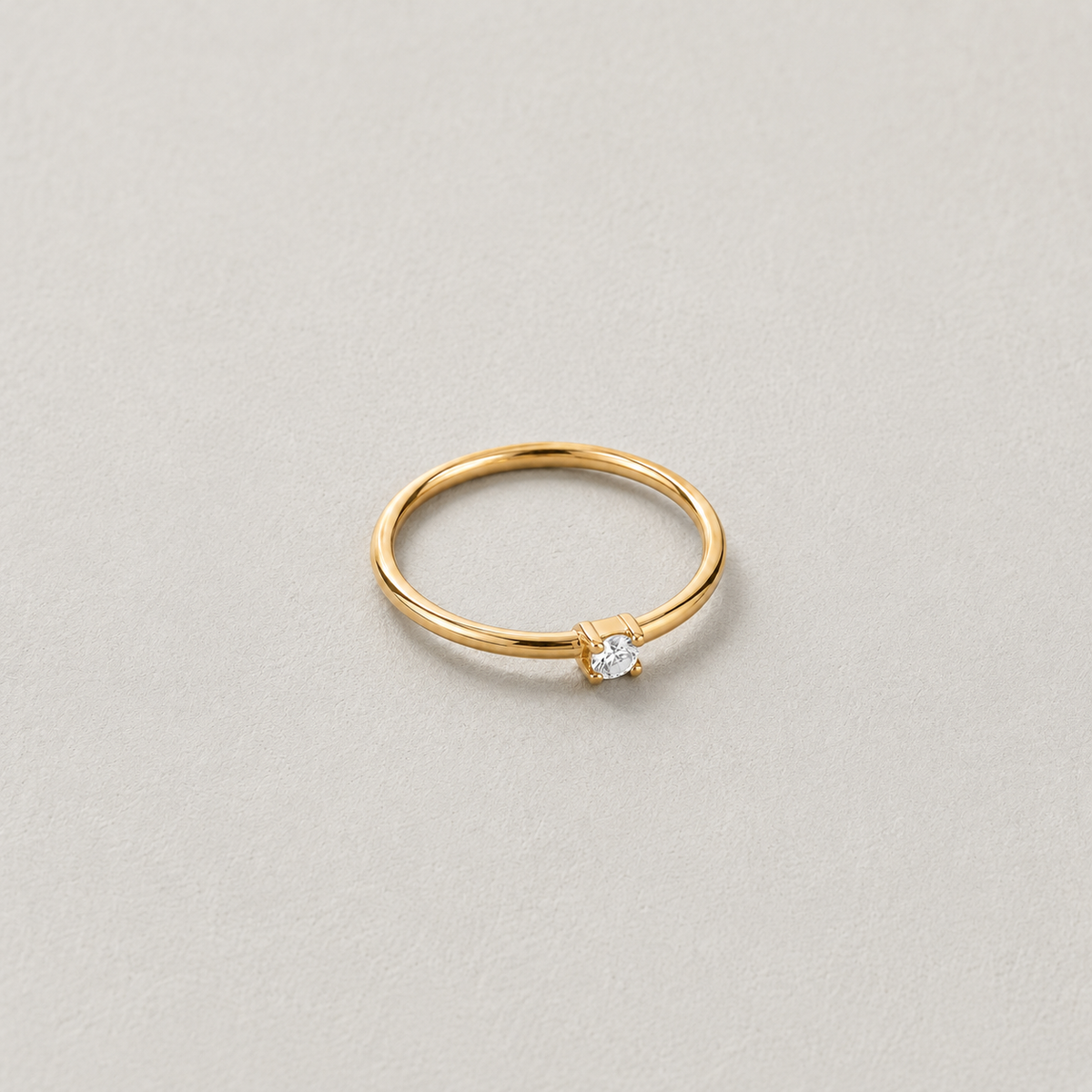 Dew Ring, smal guldfärgad ring med klar sten på ljus stenbakgrund.