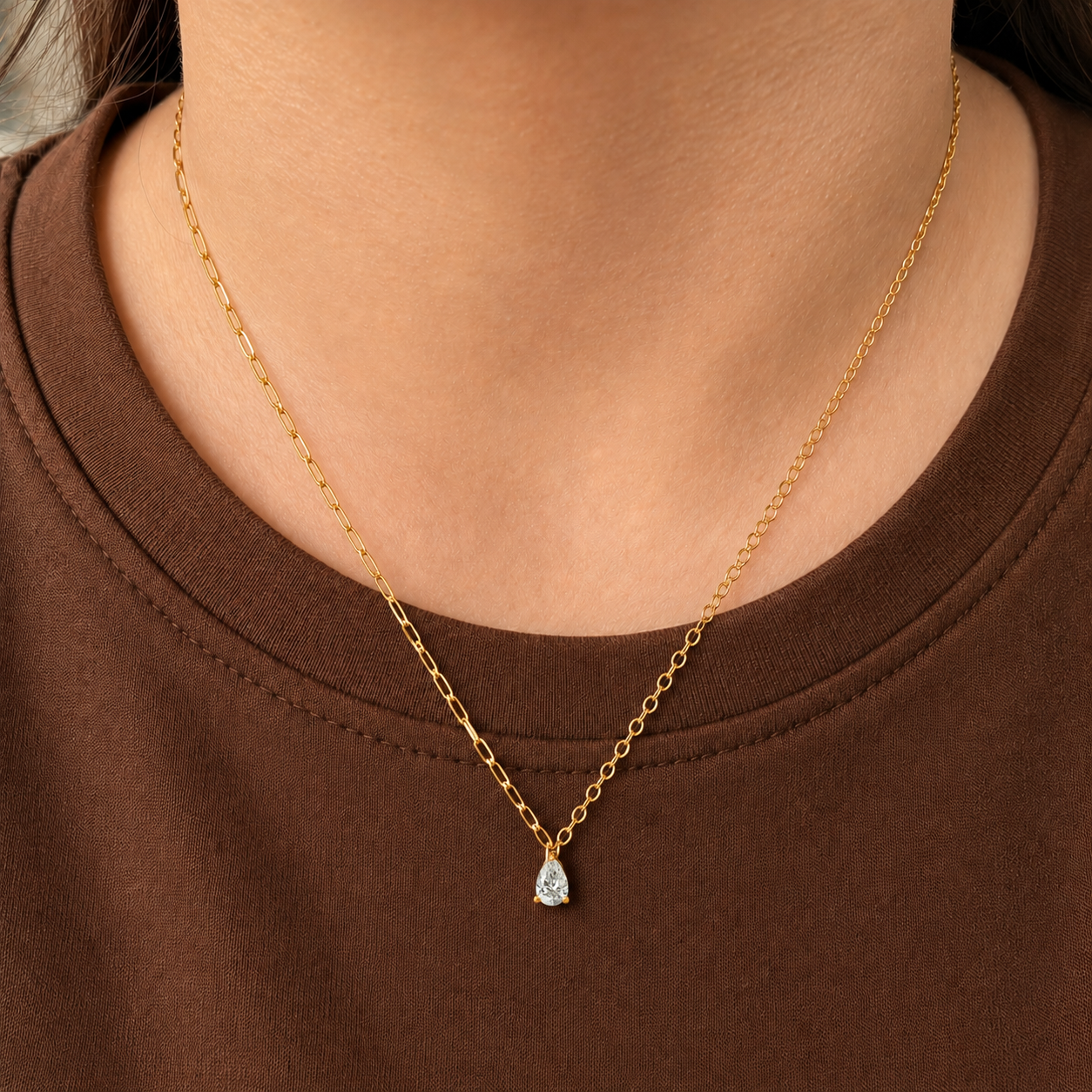 Drop Necklace i 316 kirurgiskt stål och 18k PVD-guld, ett nätt halsband med droppformat klart hänge buret på halsen mot brun textil.