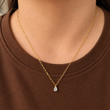 Drop Necklace i 316 kirurgiskt stål och 18k PVD-guld, ett nätt halsband med droppformat klart hänge buret på halsen mot brun textil.