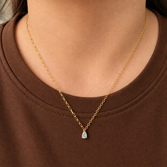 Drop Necklace i 316 kirurgiskt stål och 18k PVD-guld, ett nätt halsband med droppformat klart hänge buret på halsen mot brun textil.