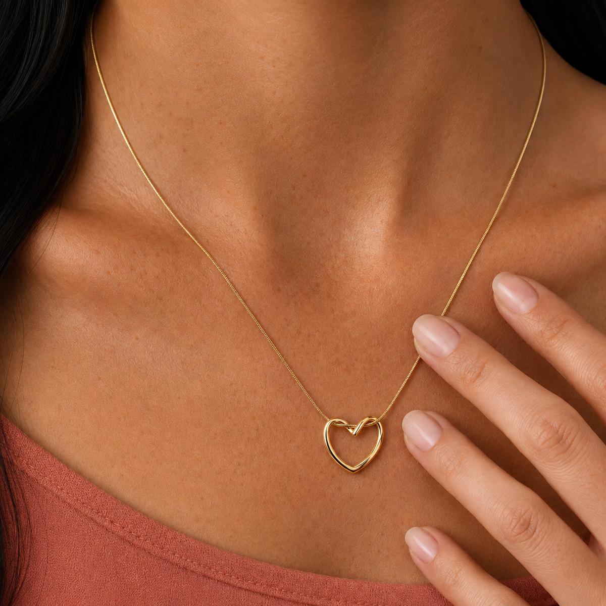 Echo Necklace i 316 kirurgiskt stål och 18k PVD-guld, guldfärgat halsband med öppet hjärthänge buret mot varm terrakottafärgad topp.