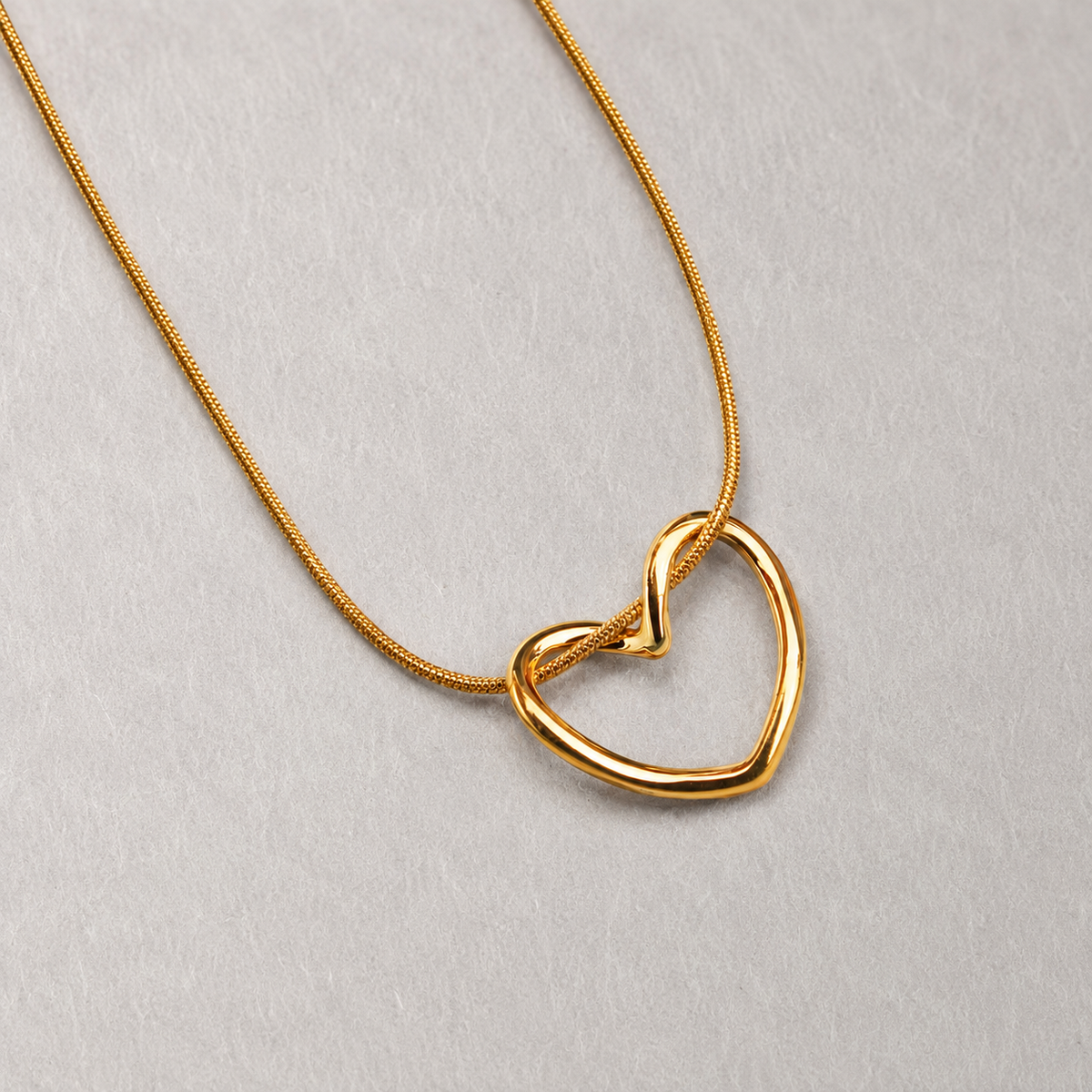 Echo Necklace i 316 kirurgiskt stål och 18k PVD-guld, guldfärgat halsband med öppet hjärthänge mot ljus neutral bakgrund.