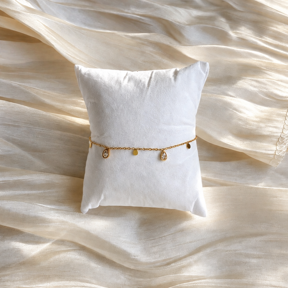 Glimmer Bracelet i 316 kirurgiskt stål och 18k PVD-guld, ett guldfärgat armband med små berlocker på vit kudde och mjuk ljus textil.