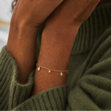 Glimmer Bracelet i 316 kirurgiskt stål och 18k PVD-guld, ett nätt armband med små berlocker buret på handleden mot grön stickad textil.