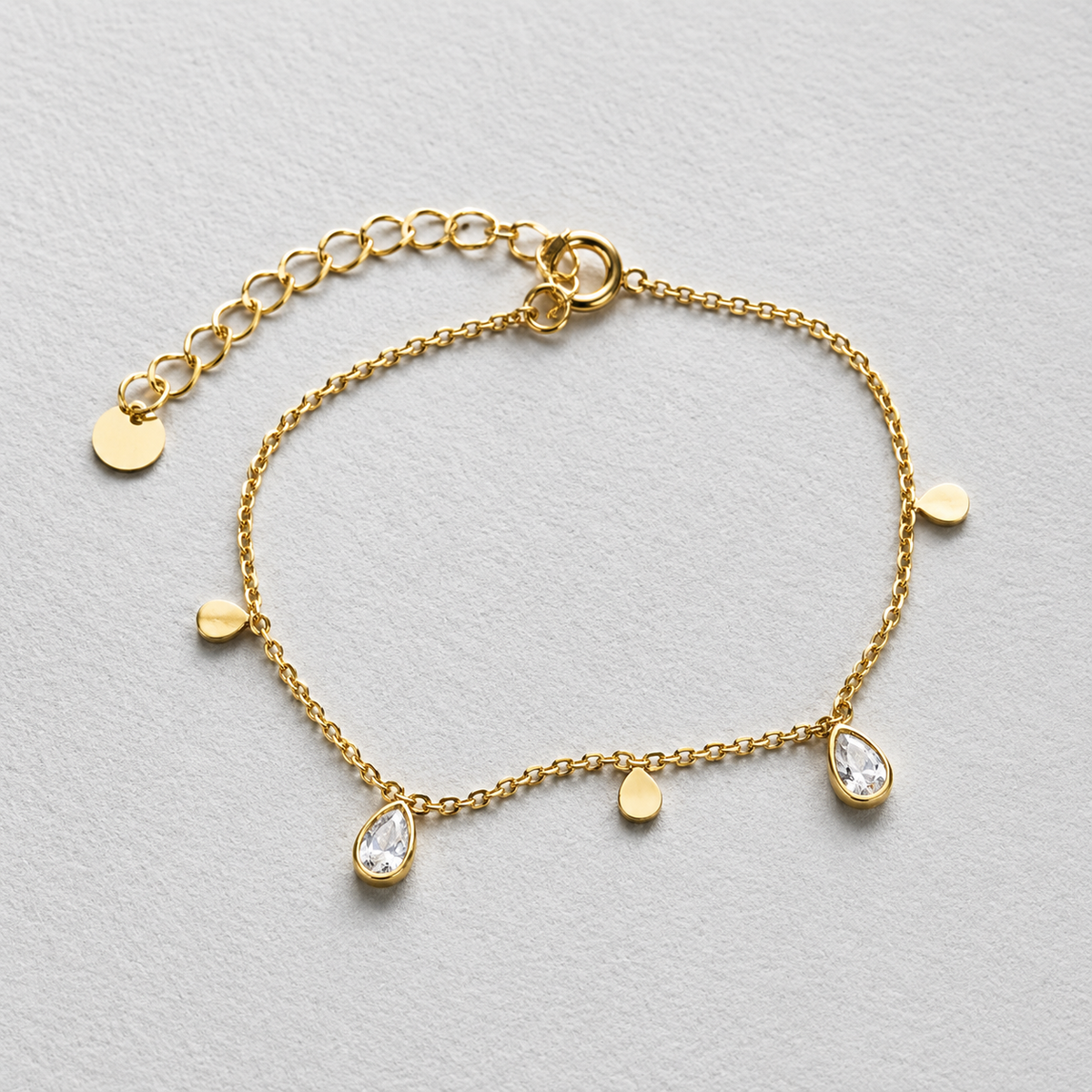 Glimmer Bracelet i 316 kirurgiskt stål och 18k PVD-guld, guldfärgat armband med små droppformade detaljer och klara stenar mot ljus neutral bakgrund.