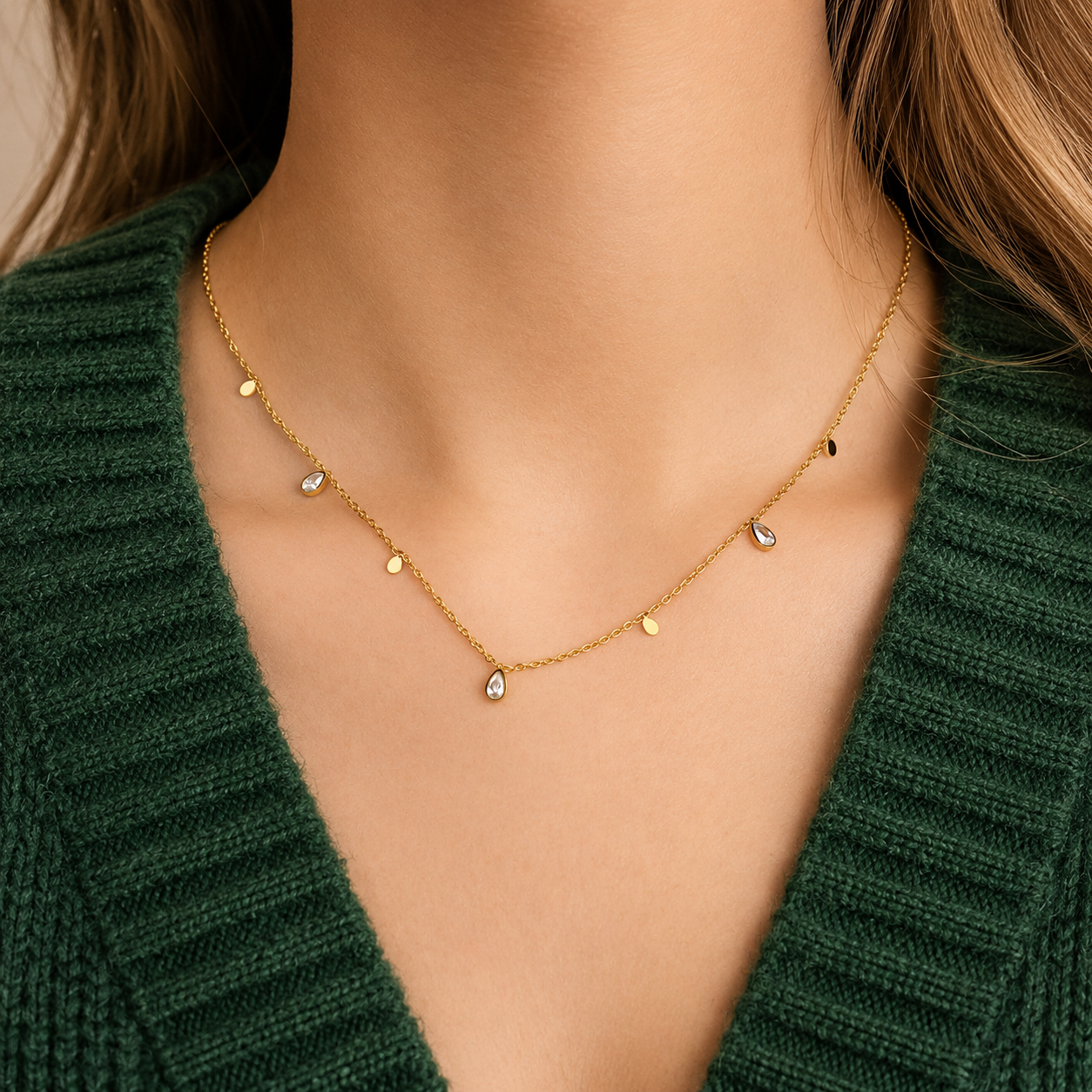 Glimmer Necklace i 316 kirurgiskt stål och 18k PVD-guld, ett nätt halsband med små berlocker buret på halsen mot mörkgrön stickad textil.