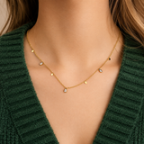 Glimmer Necklace i 316 kirurgiskt stål och 18k PVD-guld, ett nätt halsband med små berlocker buret på halsen mot mörkgrön stickad textil.