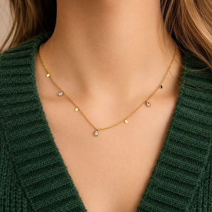 Glimmer Necklace i 316 kirurgiskt stål och 18k PVD-guld, ett nätt halsband med små berlocker buret på halsen mot mörkgrön stickad textil.