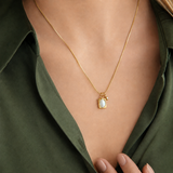 Halo Necklace i 316 kirurgiskt stål och 18k PVD-guld, ett guldfärgat halsband med ljust ovalt hänge buret på halsen mot mörkgrön skjorta.