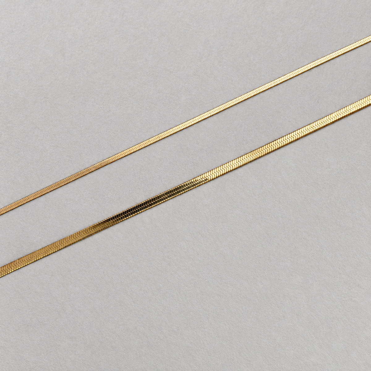 Lune Necklace 3mm och 1.7mm i guld visade som två platta kedjor på ljus stenyta.