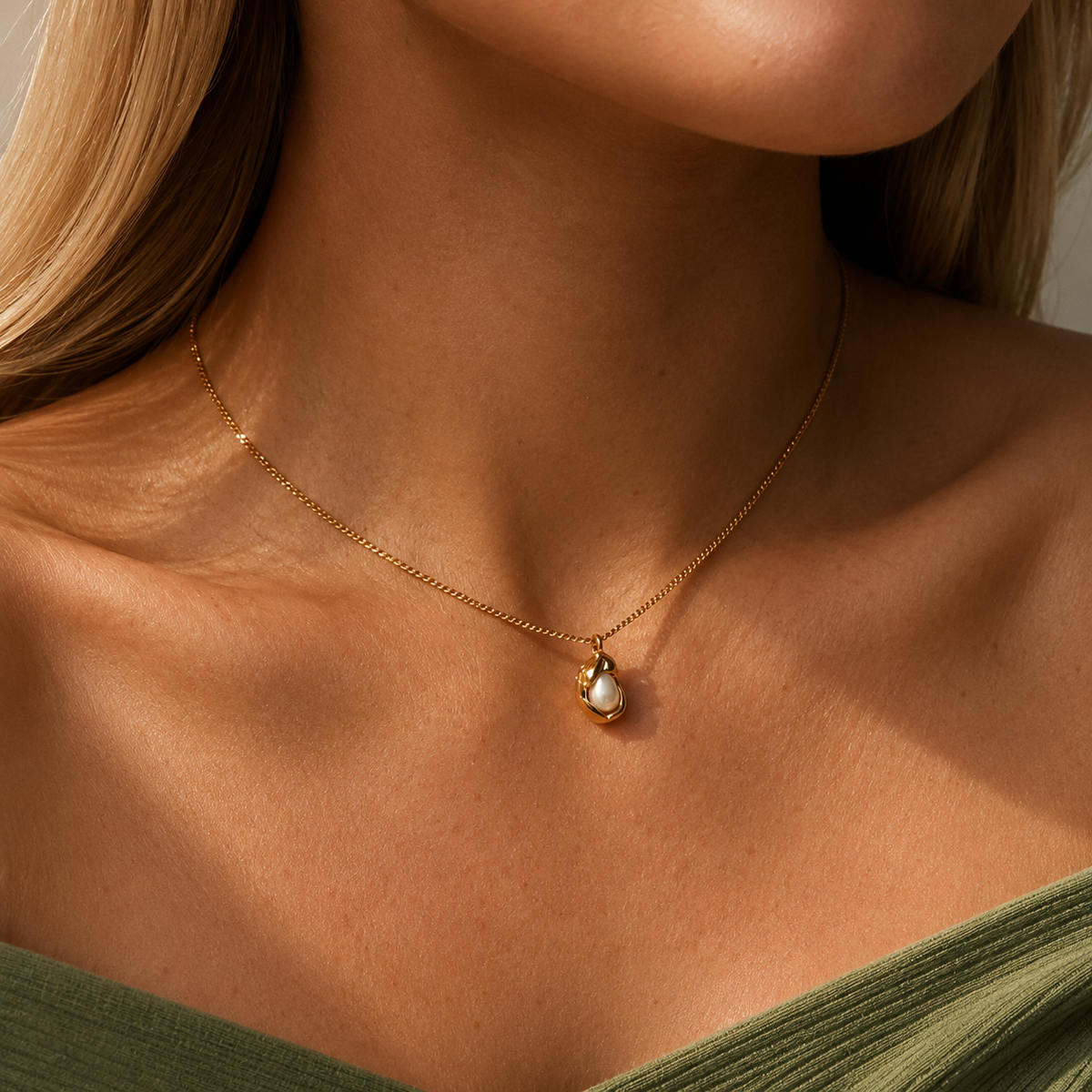 Oyster Necklace i 316 kirurgiskt stål och 18k PVD-guld, guldfärgat halsband med pärldetalj buret mot grön textil.