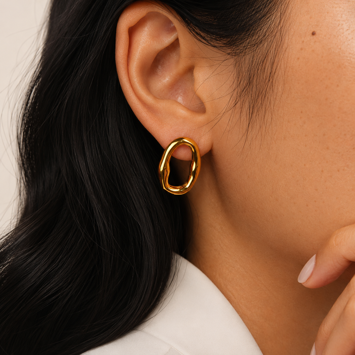 Pebble Stud i 316 kirurgiskt stål och 18k PVD-guld, ett skulpturalt ovalt örhänge buret i örat mot vit textil.