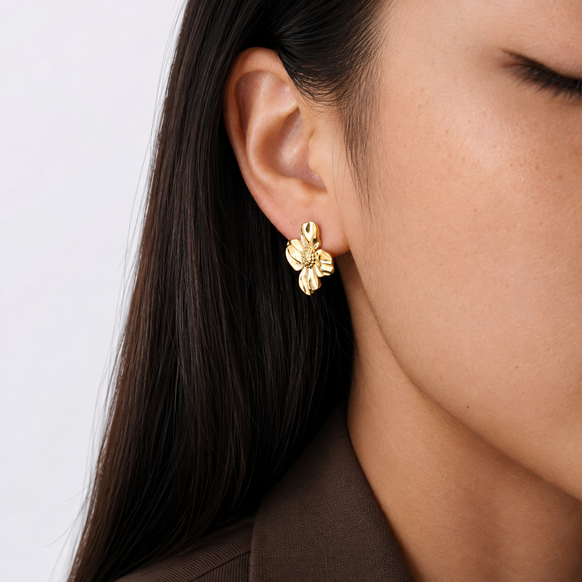Petal Stud i 316 kirurgiskt stål och 18k PVD-guld, blomformad guldfärgad stud-örhänge buren i örat mot ljus bakgrund.
