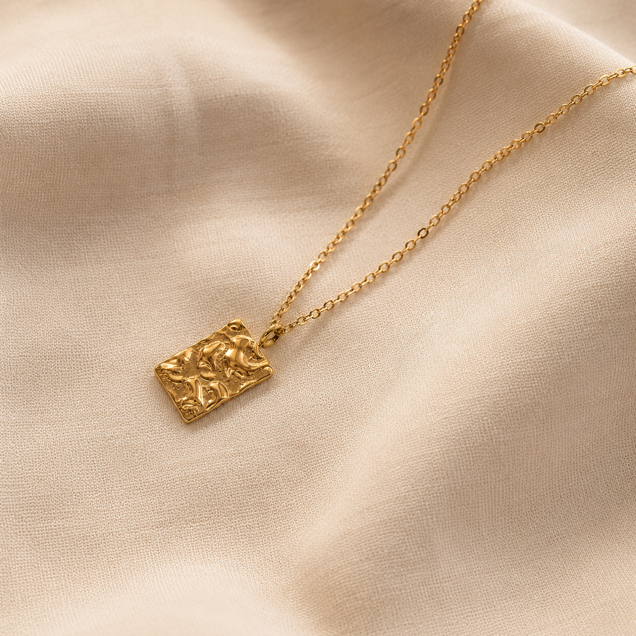 Relief Necklace i 316 kirurgiskt stål och 18k PVD-guld, ett guldfärgat halsband med texturerat rektangulärt hänge på mjuk beige textil.
