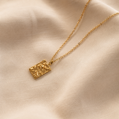 Relief Necklace i 316 kirurgiskt stål och 18k PVD-guld, ett guldfärgat halsband med texturerat rektangulärt hänge på mjuk beige textil.