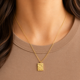 Relief Necklace i 316 kirurgiskt stål och 18k PVD-guld, ett guldfärgat halsband med texturerat rektangulärt hänge buret på halsen mot beige topp.