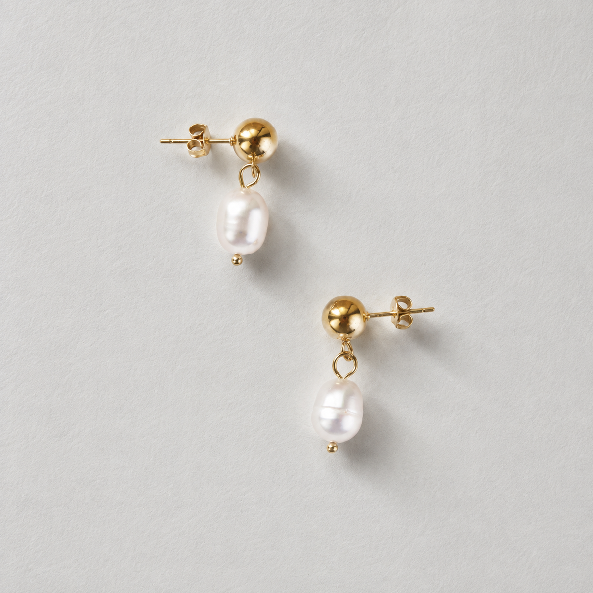 Satin Pearl Drop i 316 kirurgiskt stål och 18k PVD-guld, guldfärgade örhängen med hängande pärlor mot ljus neutral bakgrund.