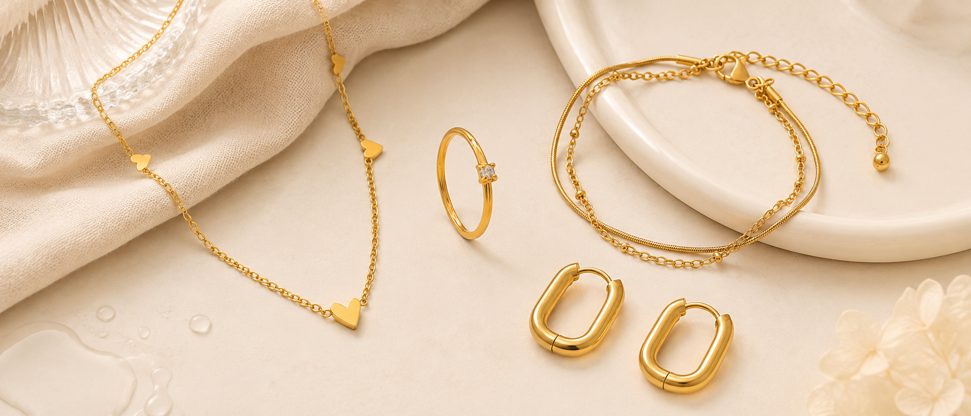 Vattentåliga smycken i 316 kirurgiskt stål och 18k PVD-guld från Serein Arc, med halsband, ring, armband och hoops på varm beige bakgrund.