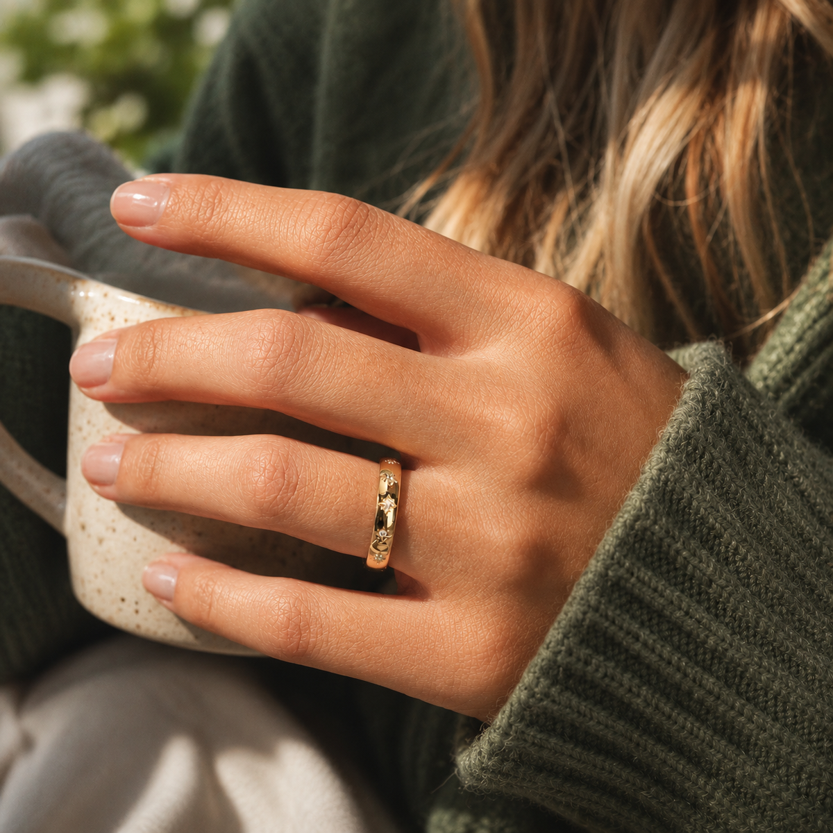 Sol Ring på hand, guldfärgad ring med små klara stenar och stjärnformade detaljer, fotograferad i varm miljö medan personen håller en keramikmugg.