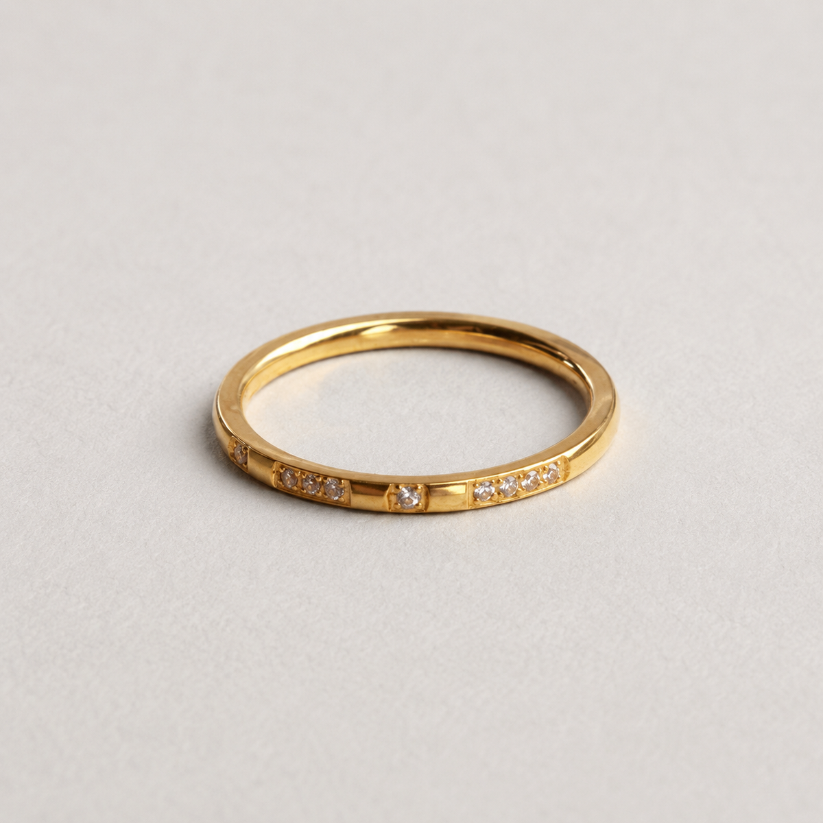 Spark Ring, smal guldfärgad ring med klara stenar mot ljus stenbakgrund.