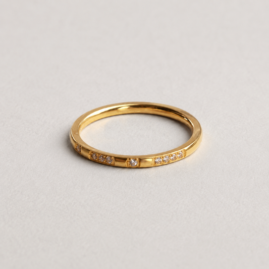 Spark Ring, smal guldfärgad ring med klara stenar mot ljus stenbakgrund.