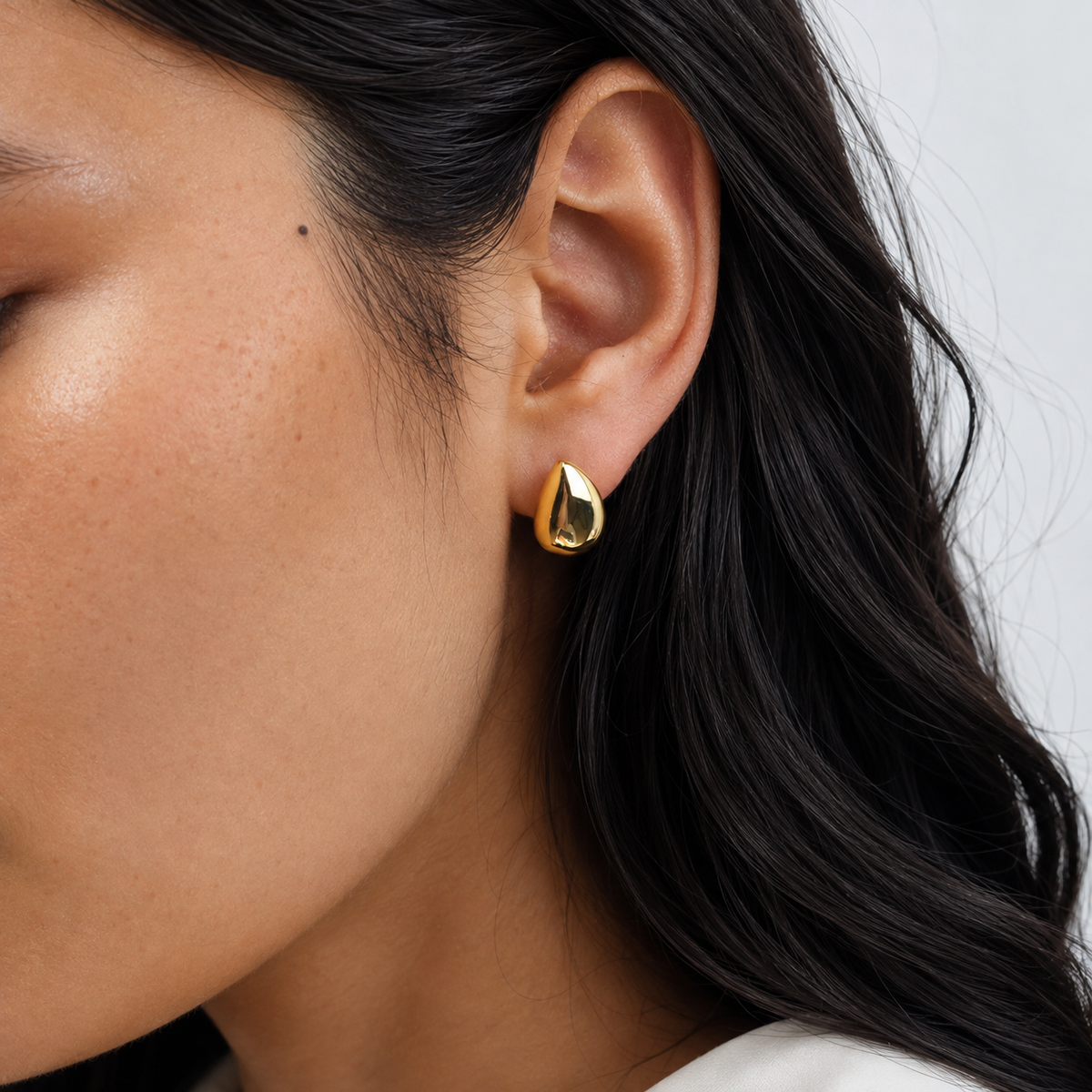 Stone Studs i 316 kirurgiskt stål och 18k PVD-guld, skulpturala droppformade örhängen burna i örat mot ljus neutral bakgrund.