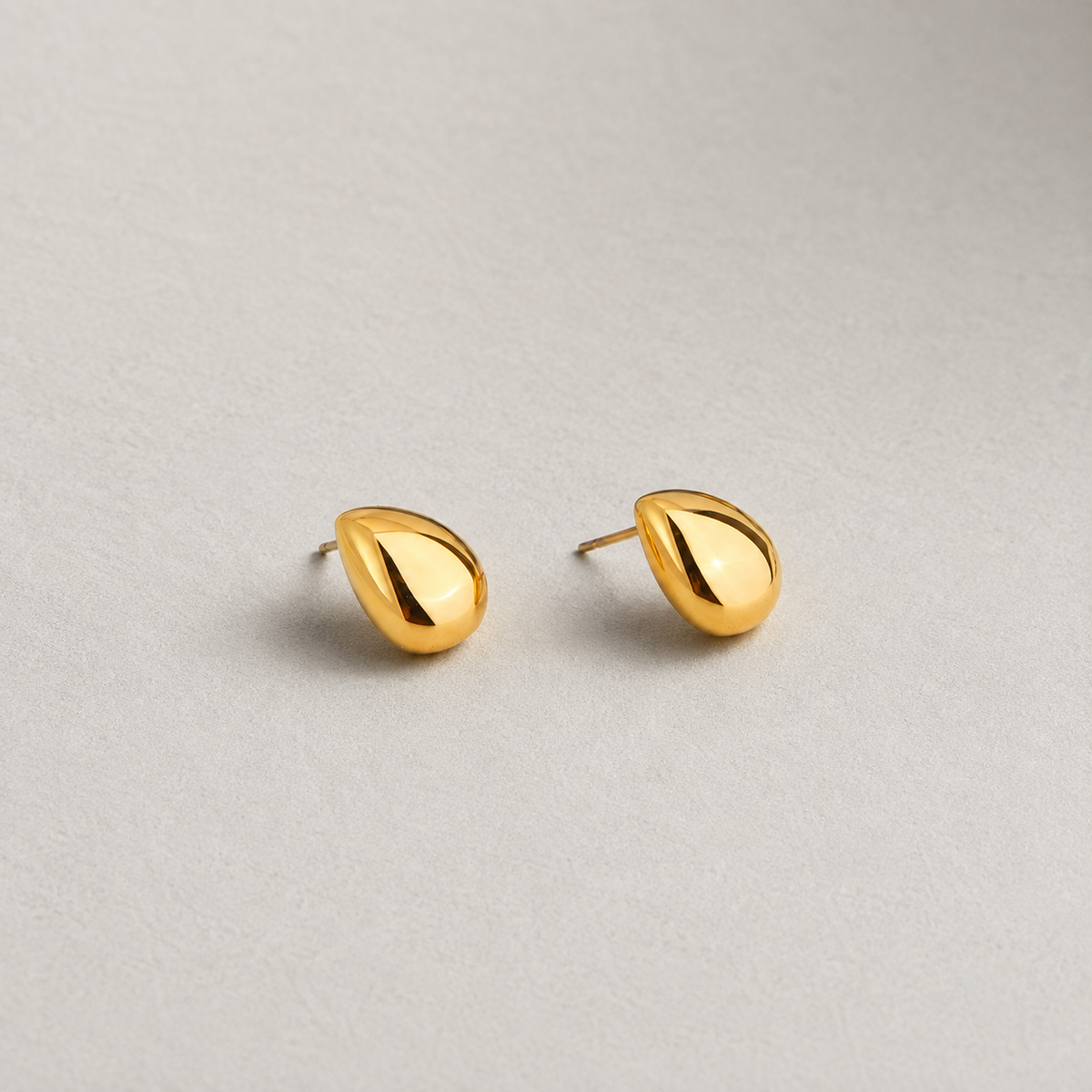 Stone Studs i 316 kirurgiskt stål och 18k PVD-guld, skulpturala droppformade örhängen mot ljus neutral bakgrund.