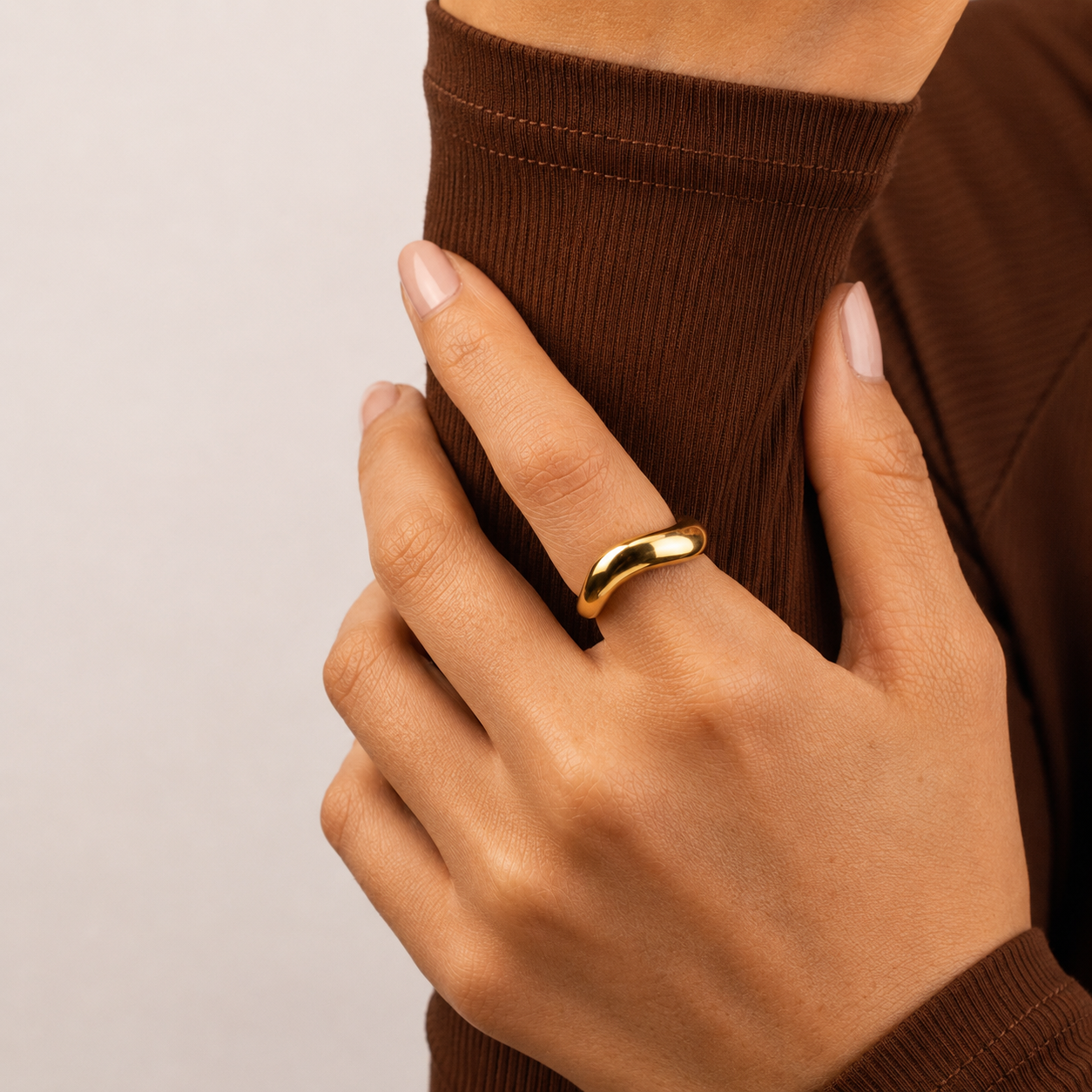 Swell Ring i 316 kirurgiskt stål och 18k PVD-guld, en mjukt rundad guldfärgad ring buren på hand mot brun textil.