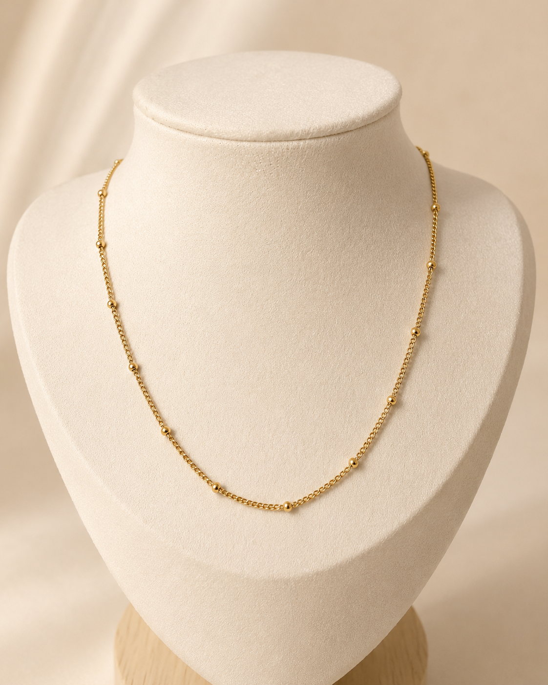 Tether Necklace i 316 kirurgiskt stål och 18k PVD-guld, ett guldfärgat halsband med små kuldetaljer på vit smyckesbyst mot varm neutral bakgrund.