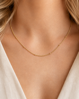 Tether Necklace i 316 kirurgiskt stål och 18k PVD-guld, ett nätt guldfärgat halsband med små kuldetaljer buret på halsen mot vit textil.
