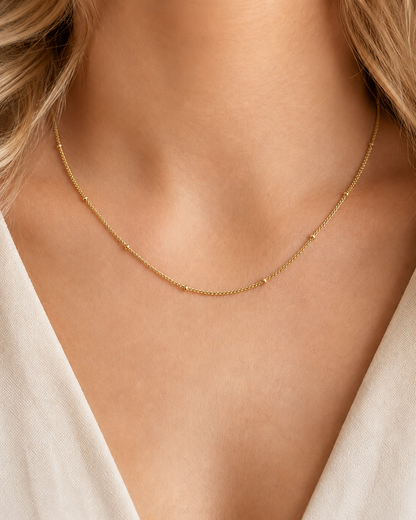 Tether Necklace i 316 kirurgiskt stål och 18k PVD-guld, ett nätt guldfärgat halsband med små kuldetaljer buret på halsen mot vit textil.