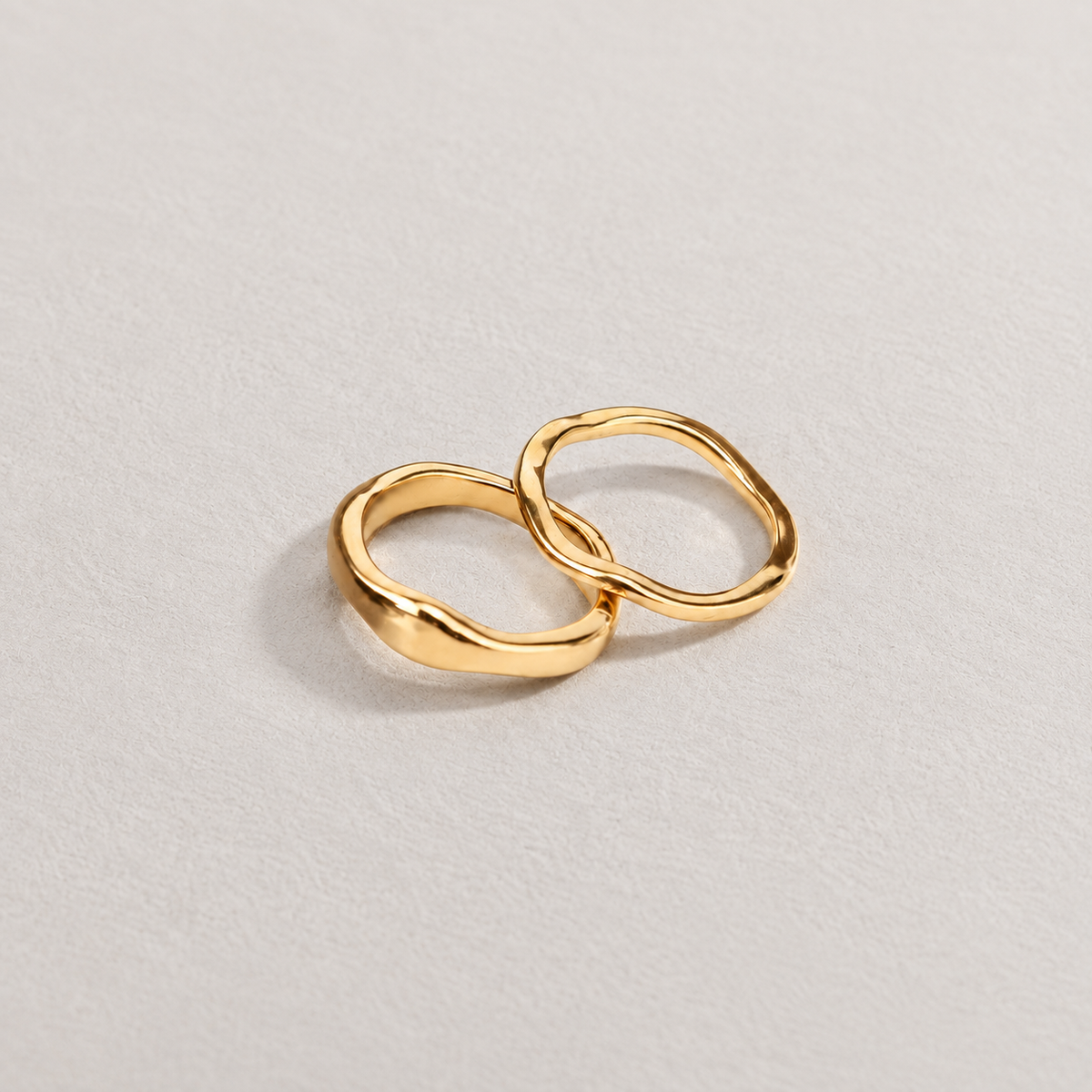 Trace Ring, två guldfärgade ringar med mjukt vågig form på ljus stenbakgrund.