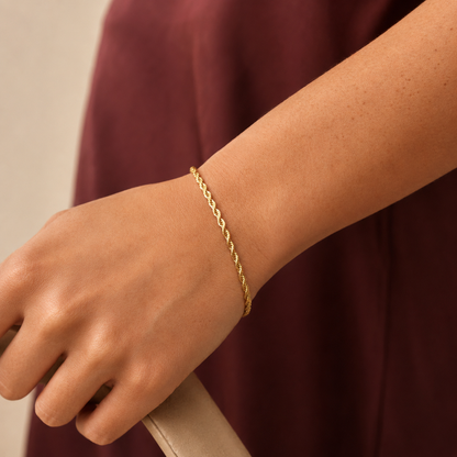 Weave Bracelet i 316 kirurgiskt stål och 18k PVD-guld, ett guldfärgat armband med tvinnad kedja buret på handleden mot vinröd textil.